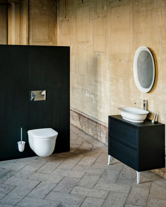 Laufen-seinäkiinnitteinen New Classic -WC-istuin. Tilassa on Laufenin keraaminen, valkoinen soikion muotoinen käsienpesuallas ja musta allaskaappi.