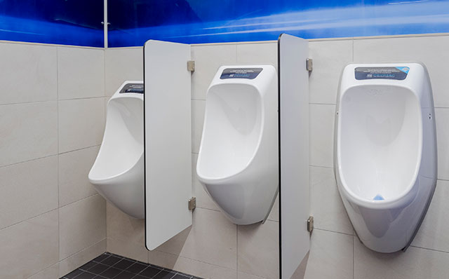 Kolme vedetöntä, valkoista urinaalia on kiinnitetty miesten vessan seinään. Urinaalit on asennettu eri korkeuksille. Urinaalien kaksi väliseinää ovat vaaleat ja eri korkeu.