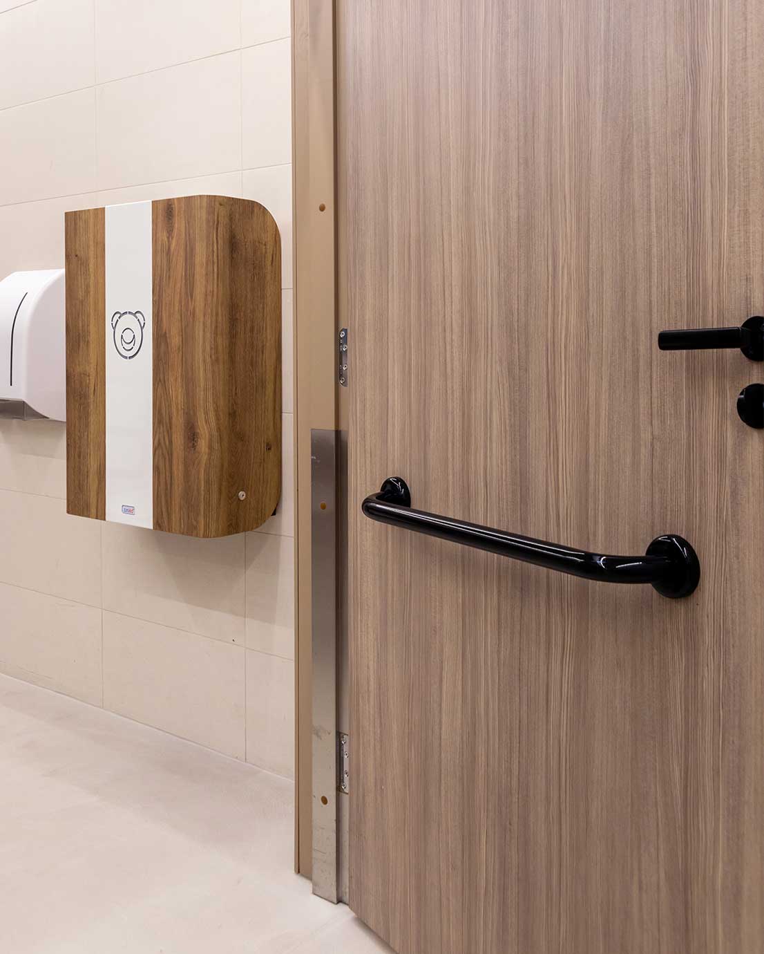 WC-tilan ovessa on puukuosinen laminaatti ja musta tukikahva. Oven vieressä seinällä on puukuosinen vaipanvaihtopöytä, jossa on valkoinen keskiosa ja nallelogo. Seinällä näkyy myös valkoinen paperiannostelija. Tausta on laatoitettu vaaleilla laatoilla.