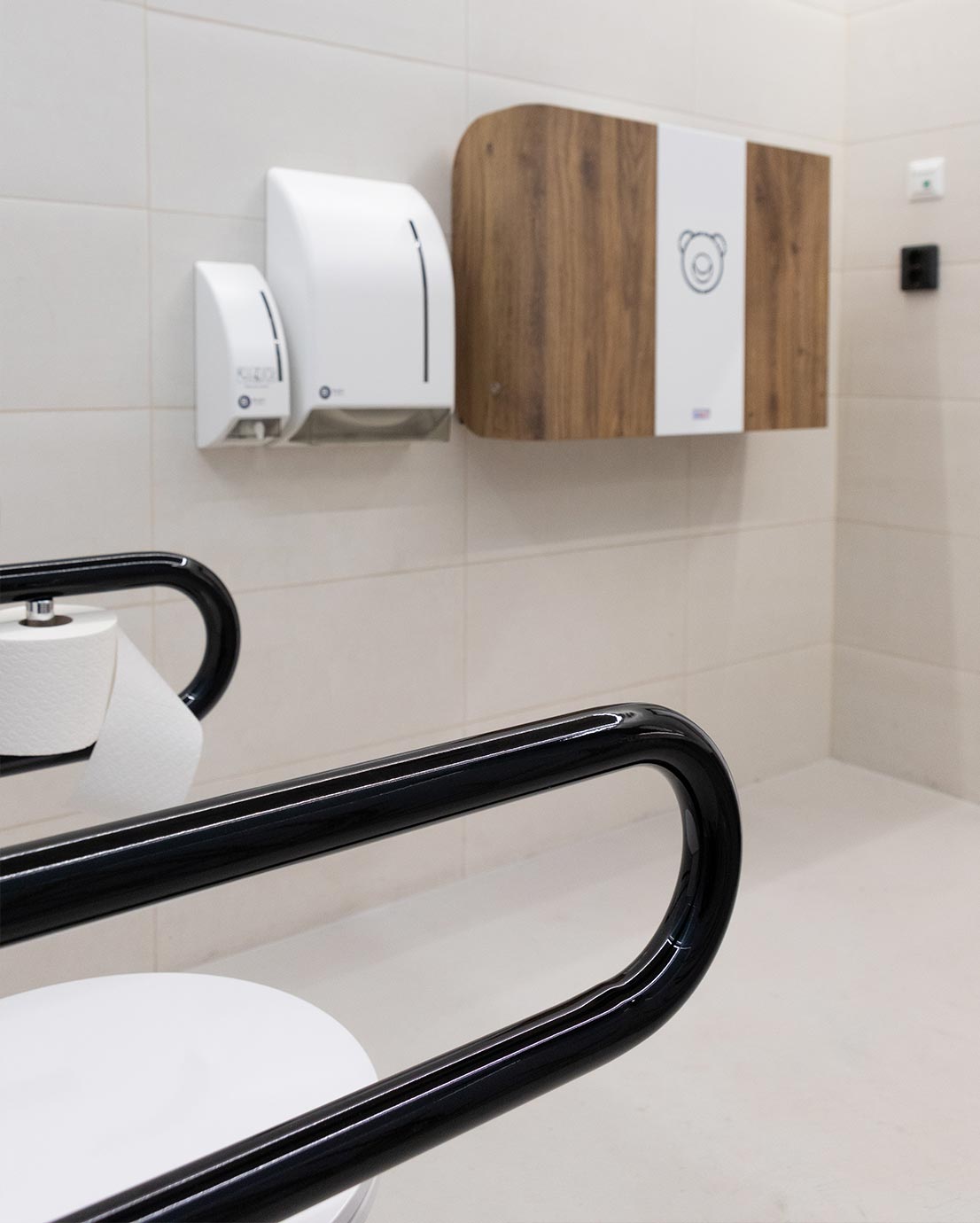 Vaaleassa esteettömässä WC-tilassa näkyvät etualalla mustat Pito-tukikahvat WC-istuimelle ja WC-paperirulla. Takaseinällä on seinään asennettava KAWAQ vauvan hoitopöytä ja BlackSatinon saippua ja -paperiannostelijat.