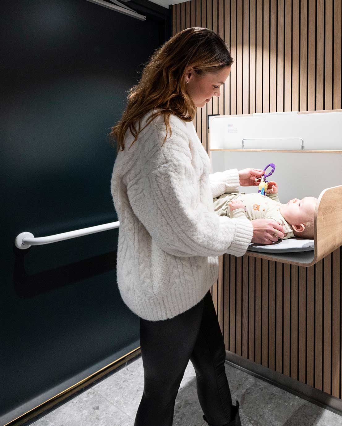 Nainen hoitaa vauvaa vaakamallisessa seinäkiinnitteisessä hoitopöydässä. Naisen takana näkyy tummanvihreä WC-tilan ovi. Ovessa on valkoinen Pito-tukikahva. Seinän materiaali: puinen rimapaneeli. Lastenhoitopöydän väri: tammi. Malli: Timkid, KAWAQ.